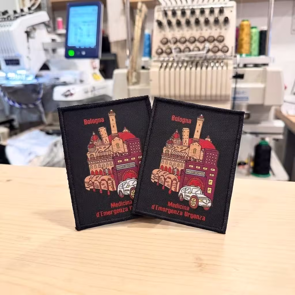 Two rectangular printed patches for Bologna emergency medicine featuring city landmarks, displayed near an embroidery machine.