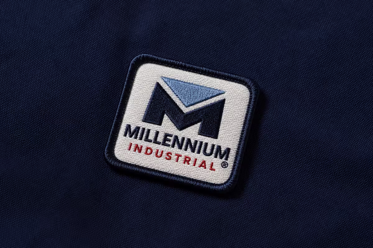 Square custom woven patch featuring the Millennium Industrial corporate logo sewn onto dark navy fabric. 