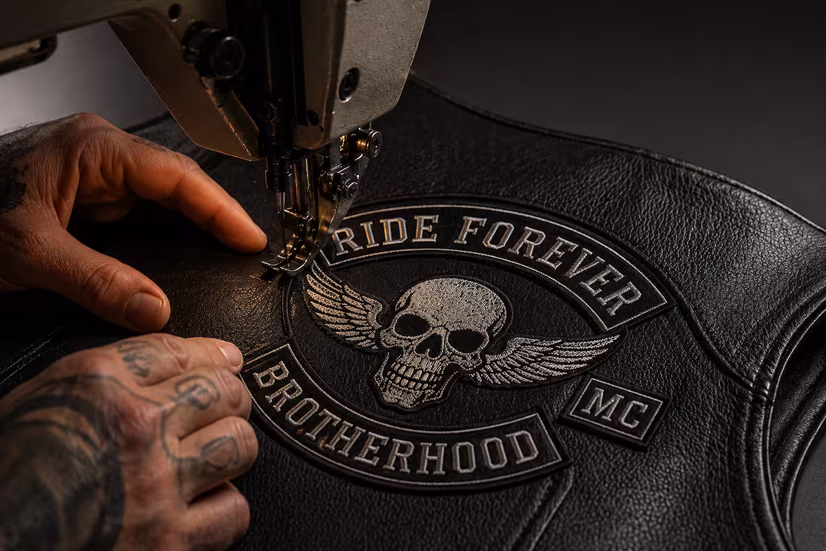 Tattooed hands using an industrial sewing machine to stitch a custom Ride Forever back patch onto a leather vest.